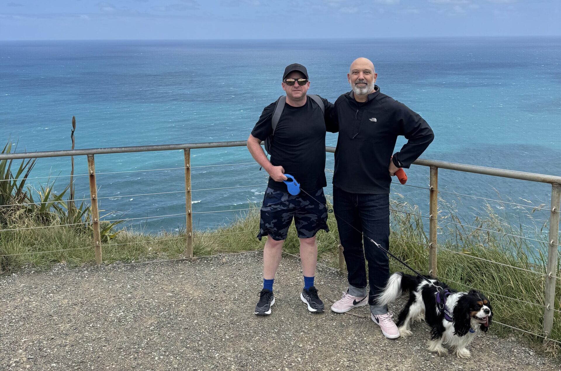 Two men standing on a path with a dog overlooking a large sea
